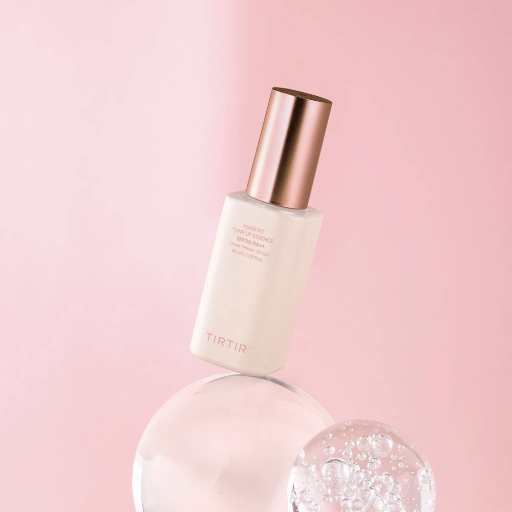 L'image montre une bouteille de "TIRTIR MASK FIT TONE UP ESSENCE" avec SPF30 PA++ sur un fond rose, ce qui donne une sensation de douceur et de féminité.