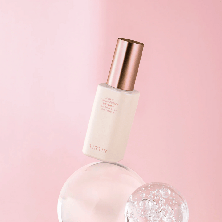 L'image montre une bouteille de "TIRTIR MASK FIT TONE UP ESSENCE" avec SPF30 PA++ sur un fond rose, ce qui donne une sensation de douceur et de féminité.