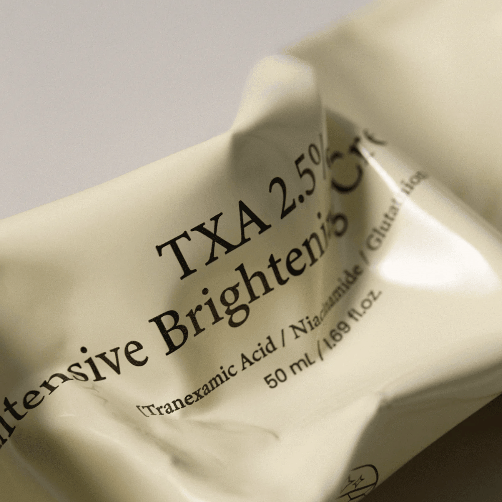 Gros plan de AXIS - Y TXA 2.5% Intensive Brightening Cream où le design doux et minimaliste du tube est au centre de l’attention. L’image met en valeur le nom du produit, l’Acide Tranexamique et la Niacinamide ainsi que l’élégant emballage beige.