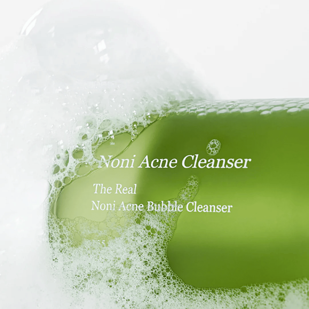 La bouteille avec The Real Noni Acne Bubble Cleanser est au centre et entourée de mousse abondante. La couleur verte fraîche signale des ingrédients naturels, parfaite pour nettoyer et combattre l'acné. Un produit idéal pour une peau propre et claire.
