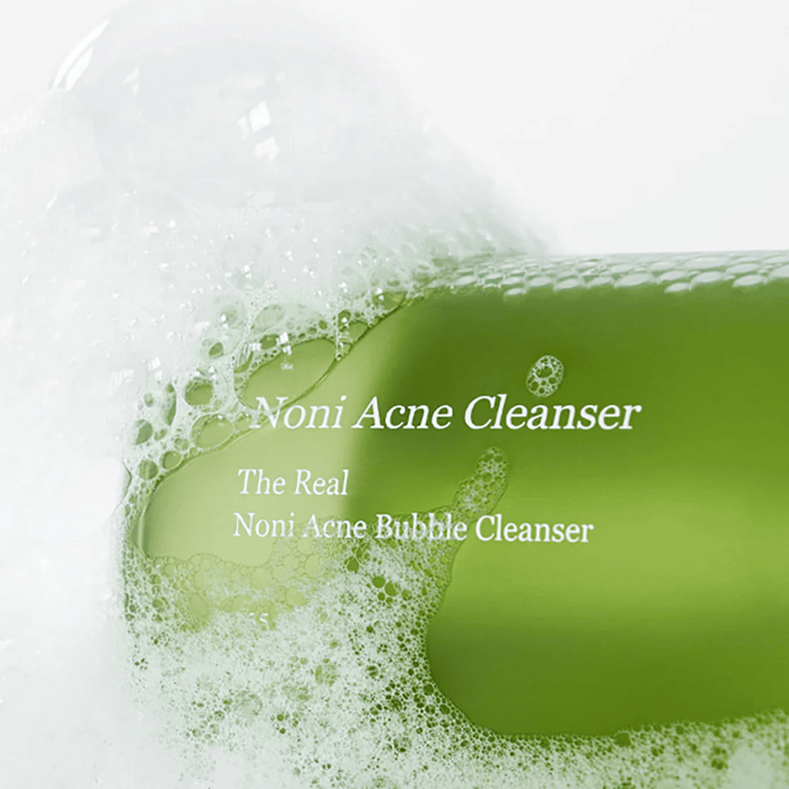 La bouteille avec The Real Noni Acne Bubble Cleanser est au centre et entourée de mousse abondante. La couleur verte fraîche signale des ingrédients naturels, parfaite pour nettoyer et combattre l'acné. Un produit idéal pour une peau propre et claire.