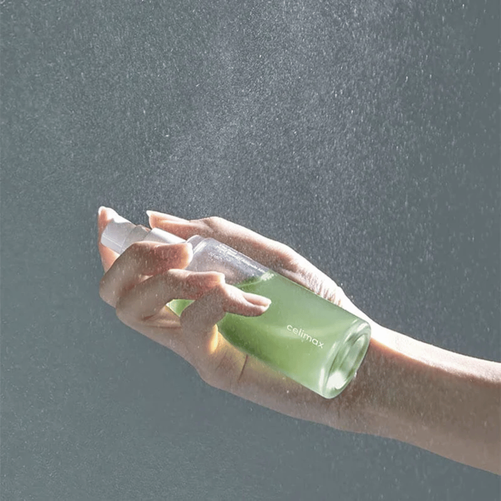 Une main tient le flacon pulvérisateur Celimax Noni Mist et applique le produit en une fine brume uniforme sur la peau. La bouteille est verte et le produit est commercialisé sous le nom de "The Real Noni Energy Ampule Mist" pour fournir de l'énergie et de l'hydratation à la peau.