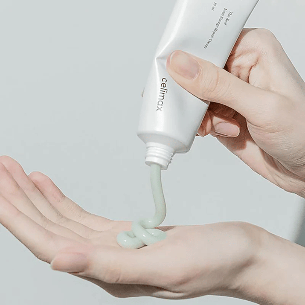 Une main tient un tube blanc avec Celimax Noni Energy Repair Cream, et la crème est pressée en un jet régulier sur la paume. La crème est vert clair et est commercialisée pour nourrir et restaurer la peau.