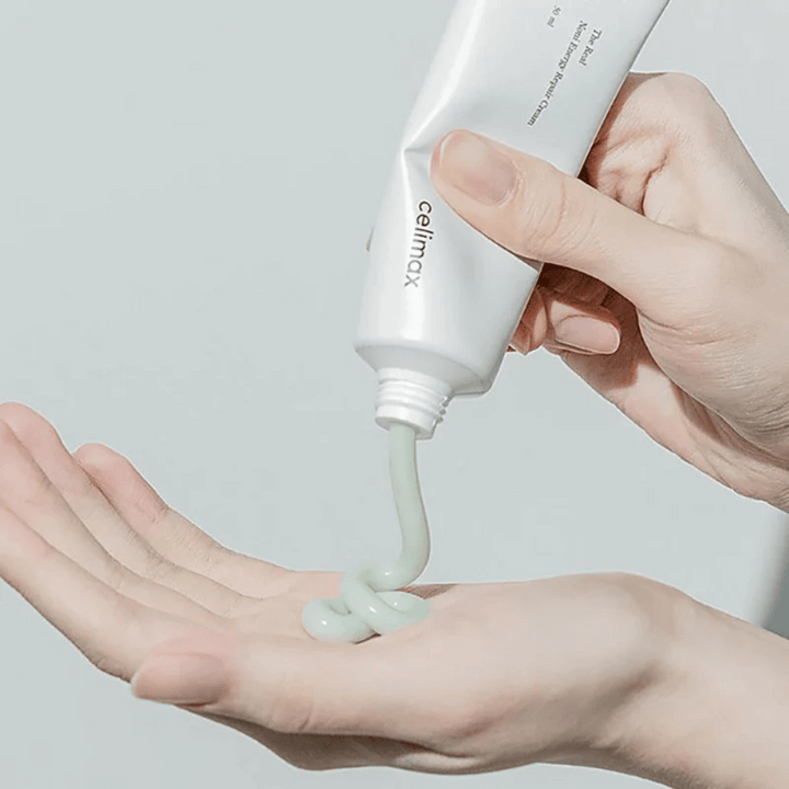 Une main tient un tube blanc avec Celimax Noni Energy Repair Cream, et la crème est pressée en un jet régulier sur la paume. La crème est vert clair et est commercialisée pour nourrir et restaurer la peau.