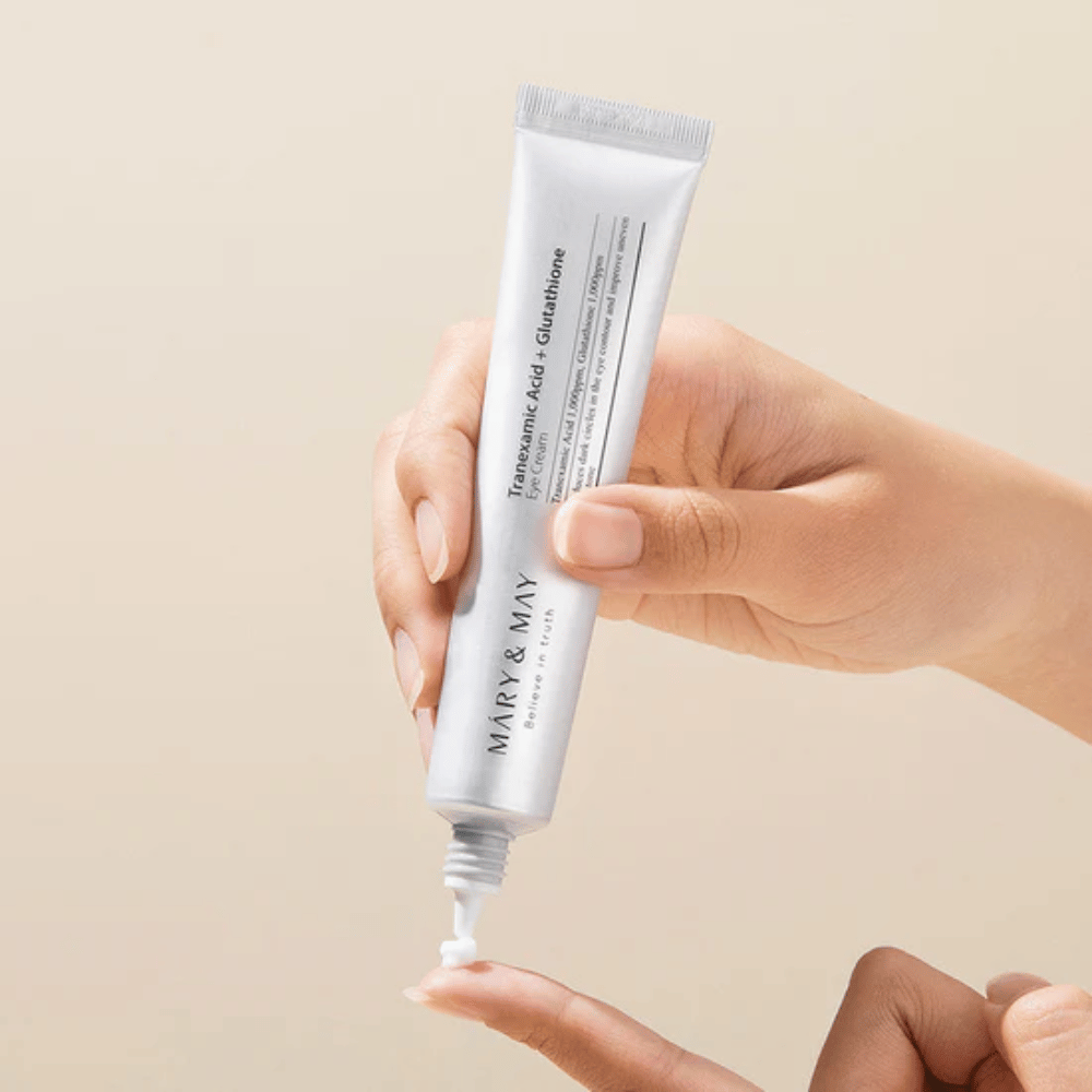 Une main tient un tube MARY & MAY "Tranexamic Acid + Glutathione Eye Cream". Le tube est blanc et le design est épuré et simple, ce qui met en valeur les propriétés apaisantes et éclaircissantes du produit pour la zone des yeux. Le produit est appliqué délicatement, avec un accent sur la précision et l'efficacité.