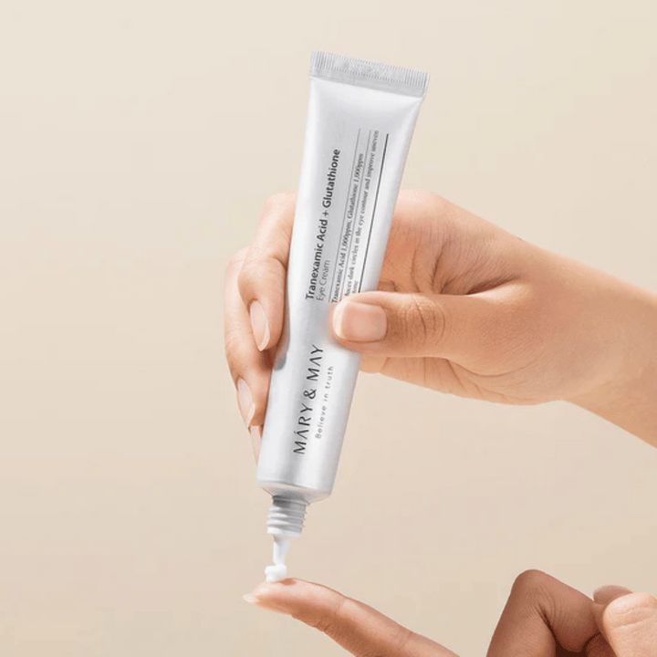 Une main tient un tube MARY & MAY "Tranexamic Acid + Glutathione Eye Cream". Le tube est blanc et le design est épuré et simple, ce qui met en valeur les propriétés apaisantes et éclaircissantes du produit pour la zone des yeux. Le produit est appliqué délicatement, avec un accent sur la précision et l'efficacité.