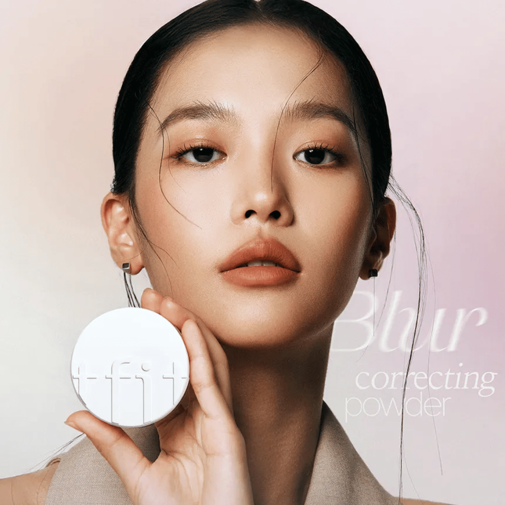 Femme avec un maquillage impeccable tenant un boîtier rond blanc avec le logo TFIT, avec le texte "Blur Correcting Powder" en arrière-plan.