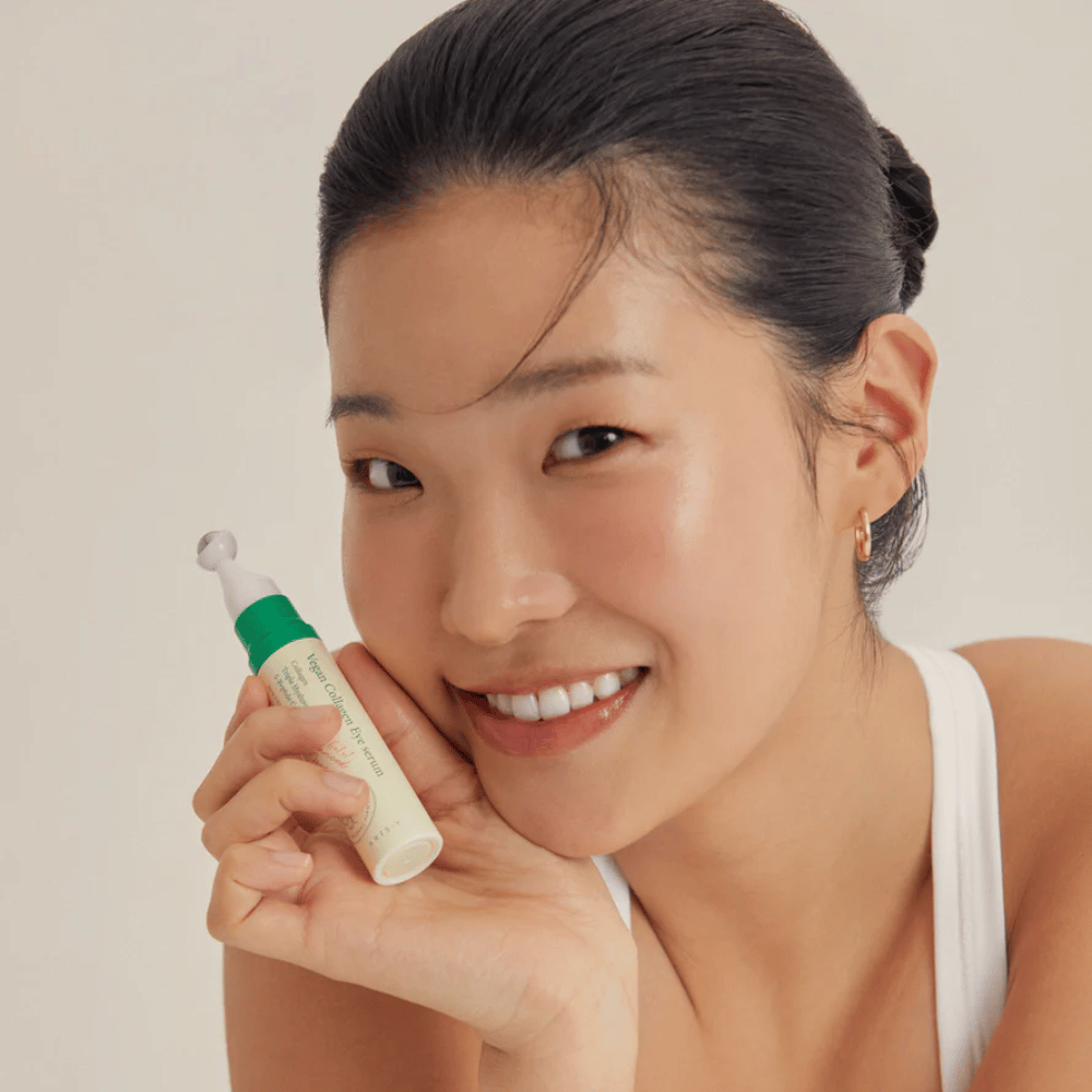 Une femme aux cheveux attachés et portant des boucles d'oreilles dorées tient un AXIS-Y Vegan Collagen Eye Serum 10ml près de son visage, tout en souriant doucement. Le sérum est conçu pour hydrater et réduire les rides autour des yeux, spécialement formulé pour les peaux sensibles.