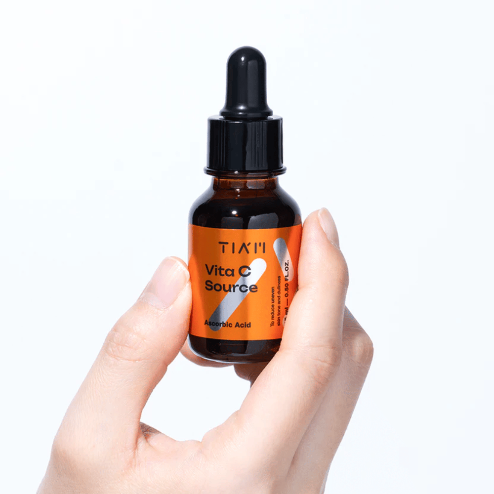 Main tenant TIA'M Vita C Source, un sérum à la vitamine C dans un flacon en verre brun avec pipette. Le produit est conçu pour uniformiser le teint et donner de l'éclat.