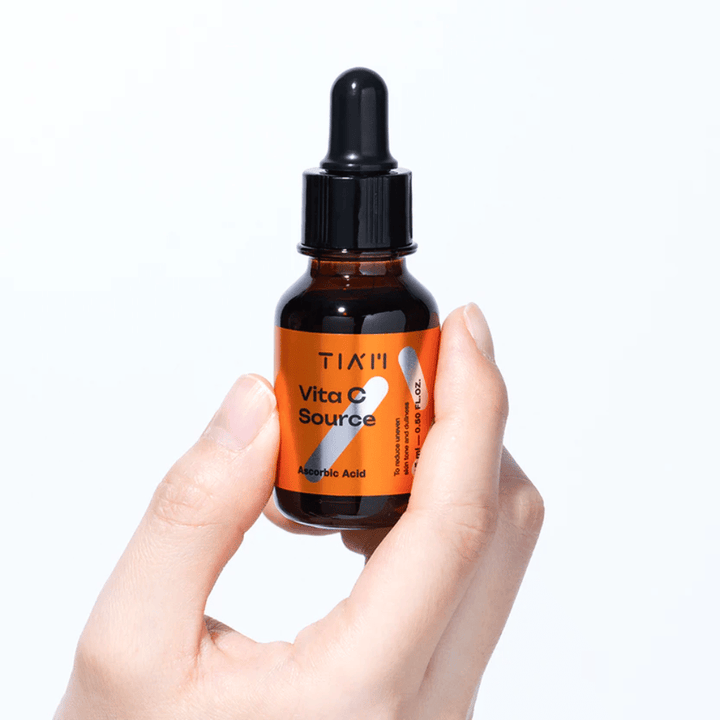 Main tenant TIA'M Vita C Source, un sérum à la vitamine C dans un flacon en verre brun avec pipette. Le produit est conçu pour uniformiser le teint et donner de l'éclat.