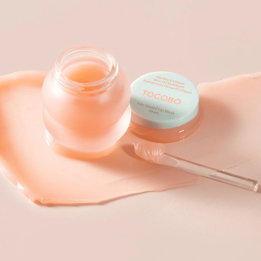 Un pot avec "Vita Glazed Lip Mask" de Tocobo est présenté. Le pot est transparent avec un couvercle turquoise clair et contient un masque à lèvres brillant et rose. Une petite spatule est placée à côté du pot, et une partie du masque est étalée sur la surface. Le produit est conçu pour hydrater et nourrir les lèvres.