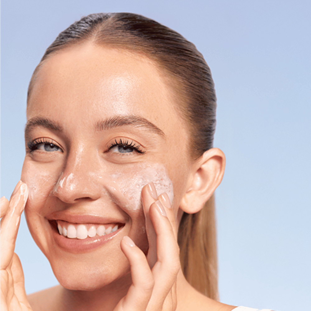 Femme souriante applique LANEIGE Water Bank Gentle Gel Cleanser sous forme moussante sur les joues, sur fond bleu.