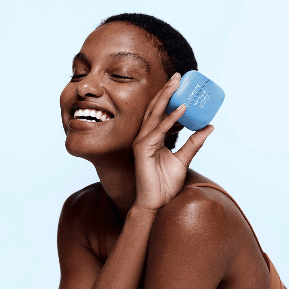 Femme souriante tenant un pot bleu LANEIGE Water Bank Blue Hyaluronic Cream contre sa joue, avec un fond bleu clair.