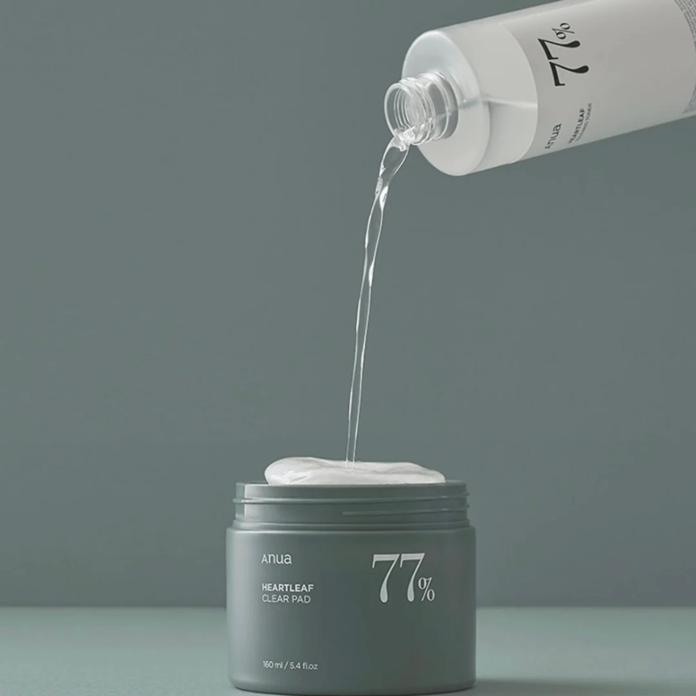 Une bouteille transparente verse un liquide dans un pot gris marqué "anua HEARTLEAF 77% Clear Pad", qui contient une pile de tampons de soin de la peau blancs. Le fond est simple et uni dans une douce teinte grise.