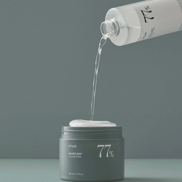 Une bouteille transparente verse un liquide dans un pot gris marqué "anua HEARTLEAF 77% Clear Pad", qui contient une pile de tampons de soin de la peau blancs. Le fond est simple et uni dans une douce teinte grise.