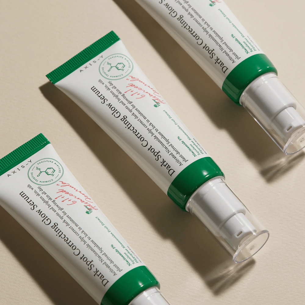 Trois tubes de Axis-Y Dark Spot Correcting Glow Serum avec bouchon vert et emballage blanc sont posés sur une surface claire.