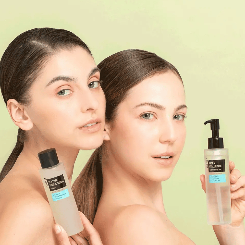 Deux femmes aux épaules dénudées posent avec des produits de soins de la peau; l'une tient une bouteille étiquetée "TEA TREE PORE & SEBUM", et l'autre un flacon pompe étiqueté "ULTRA HYALURONIC CLEANSING OIL". Elles se tiennent devant un fond vert clair.