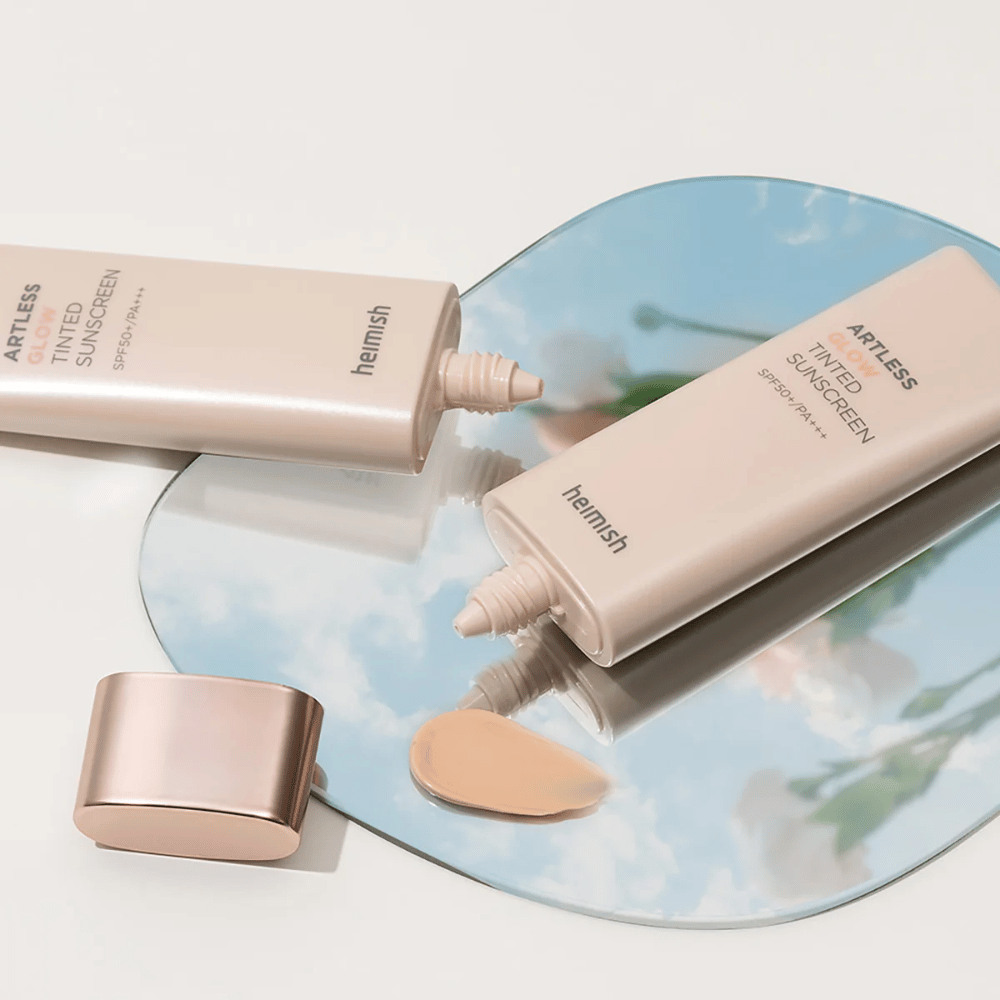 Une bouteille heimish Artless Glow Tinted Sunscreen SPF50+ PA+++ 40ml et une bouteille concealer sur un miroir, montrant différentes teintes de peau.