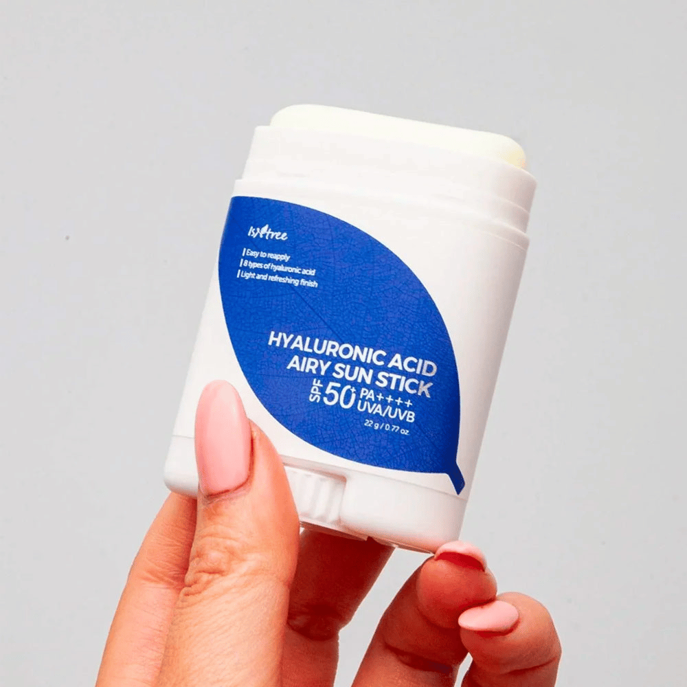 Une main tient un bâton solaire blanc avec une étiquette bleue de la marque Isntree. Le produit s'appelle "Hyaluronic Acid Airy Sun Stick" et a un facteur de protection solaire SPF 50+ PA++++.