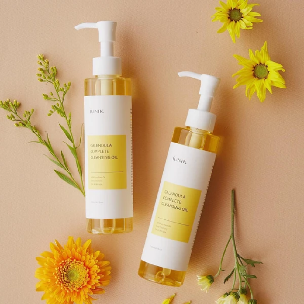 Deux bouteilles iUNIK Calendula Complete Cleansing Oil 200ml avec des fleurs jaunes sur fond beige, infusées avec de l'huile végétale.