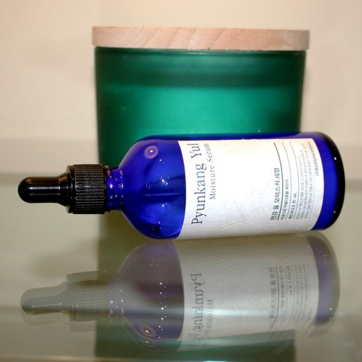 Une bouteille de Pyunkang Yul Moisture Serum 100ml équilibre les niveaux d'hydratation sur une table à côté d'une bougie.