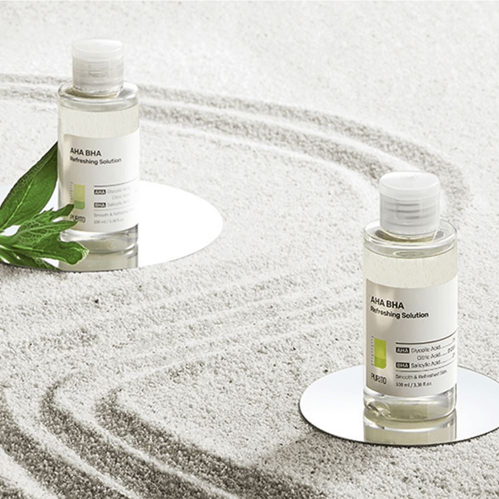 Deux PURITO AHA BHA Refreshing Solution bouteilles de 100 ml posées sur une surface sablonneuse.